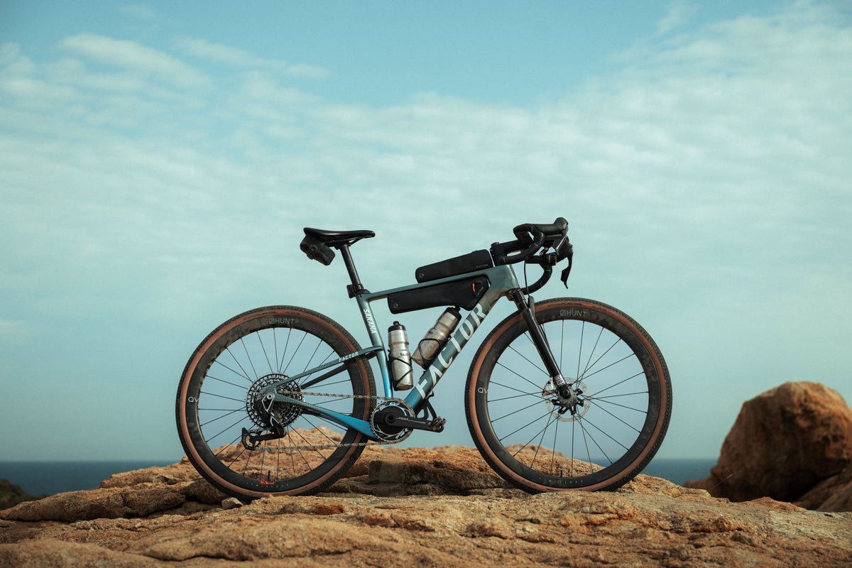 A gravel bicycle with a black frame and blue accents is parked on a rocky outcrop with the ocean and sky in the background. The bike is equipped with a black top tube bag, a water bottle, and a rear rack bag.